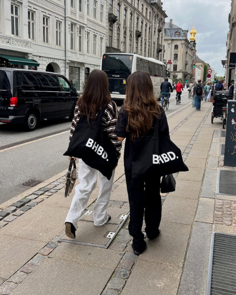 Two people walking on a city street carrying BHBD Tote Bags – black cotton tote bags with white BHBD logo, durable and lightweight for everyday use
