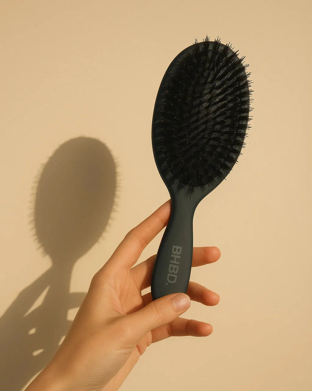 BHBD Paddle Brush – hand holding a black paddle hairbrush with natural boar and nylon bristles for smoothing and detangling hair against a beige background
